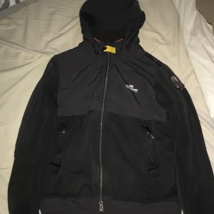 Svart jacka från Parajumpers - Säljer en svart Parajumpers jacka i mycket bra skick. Den har en huva och dragkedja framtill, samt praktiska fickor med dragkedjor. Perfekt för höst och vår, med en stilren design som funkar till det mesta. Passar dig som gillar en normal passform.