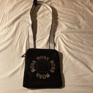 Svart axelväska från Boss - Snygg svart axelväska från Boss med ett coolt tryck av märkesnamnet i silver på framsidan. Väskan har en justerbar axelrem i vitt och svart, perfekt för vardagsbruk. Den har en dragkedja upptill för säker förvaring av dina saker. Perfekt för att ge din outfit en stilren touch!