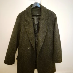 Mörkgrå kappa - Säljer en stilren mörkgrå kappa i ullblandning. Perfekt för höst och vinter. Kappan har en snygg krage och knäppning framtill. Passar både till vardags och festliga tillfällen.