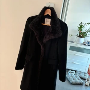 Svart kappa med pälsdetaljer - Säljer en elegant svart kappa med mjuka pälsdetaljer längs kragen. Perfekt för kyliga höst- och vinterdagar. Den har en klassisk design med långa ärmar och en rak passform. Ett måste för garderoben om du vill hålla stilen i kylan! 🖤