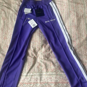 Palm Angels Byxor Lila - Säljer mina Palm Angels Byxor i en fantastisk lila färg. Byxorna är äkta och autentifierade av Vestiaire Collectives. De är använda endast en gång och är som helt nya. För fler frågor, bilder eller bud hör gärna av er!