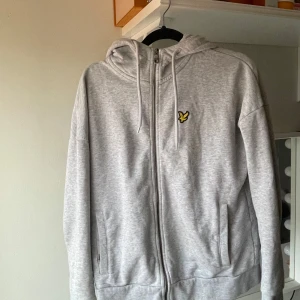 Grå hoodie från Lyle & Scott - Säljer en snygg och bekväm grå hoodie från Lyle & Scott. Den har en dragkedja framtill och en klassisk logga på bröstet. Den är i mycket bra skick och passar perfekt.