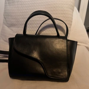ATP Atelier arezzo black Leather handbag - Säljer en elegant svart handväska i veganskt läder med stilren design. Väskan har en unik asymmetrisk flik och silverfärgade metalldetaljer. Säljer endast då den ej kommer till någon användning längre. 