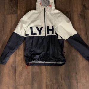 Svart och vit vindjacka från Helly Hansen - Säljer en snygg svart och vit vindjacka från Helly Hansen. Jackan har en dragkedja framtill och en justerbar huva med orange detaljer. Perfekt för blåsiga dagar och har en cool, sportig look med stora bokstäver över bröstet.