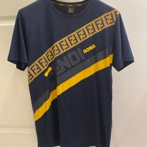 Fendi Roma T-shirt  - Snygg blå t-shirt från Fendi med gula detaljer samt ”diamanter” på trycket. Den har ett coolt mönster med Fendi-loggan och en gul rand över bröstet. På baksidan finns ett tryck med Fendi loggan. 