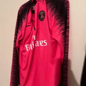 Säljer en snygg rosa fotbollströja från Paris Saint-Germain med svart mönster på ärmarna. Tröjan har en halv dragkedja och klubbens emblem på bröstet. Perfekt för träning eller som en del av din samling!