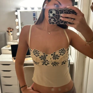 Beige korsettopp med spetsdetaljer - Säljer en superfin beige korsettopp med vackra spetsdetaljer i svart och blommönster. Toppen har justerbara axelband och en tight passform som ger en smickrande siluett. Perfekt för en utekväll eller fest! 🖤