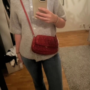 Röd axelväska i syntet - Säljer en superfin röd axelväska i syntetmaterial. Väskan har ett snyggt rynkat mönster och en praktisk axelrem. Perfekt för både vardag och fest! Den har flera fack inuti för att organisera dina saker. Väskan är i mycket bra skick och redo för en ny ägare.