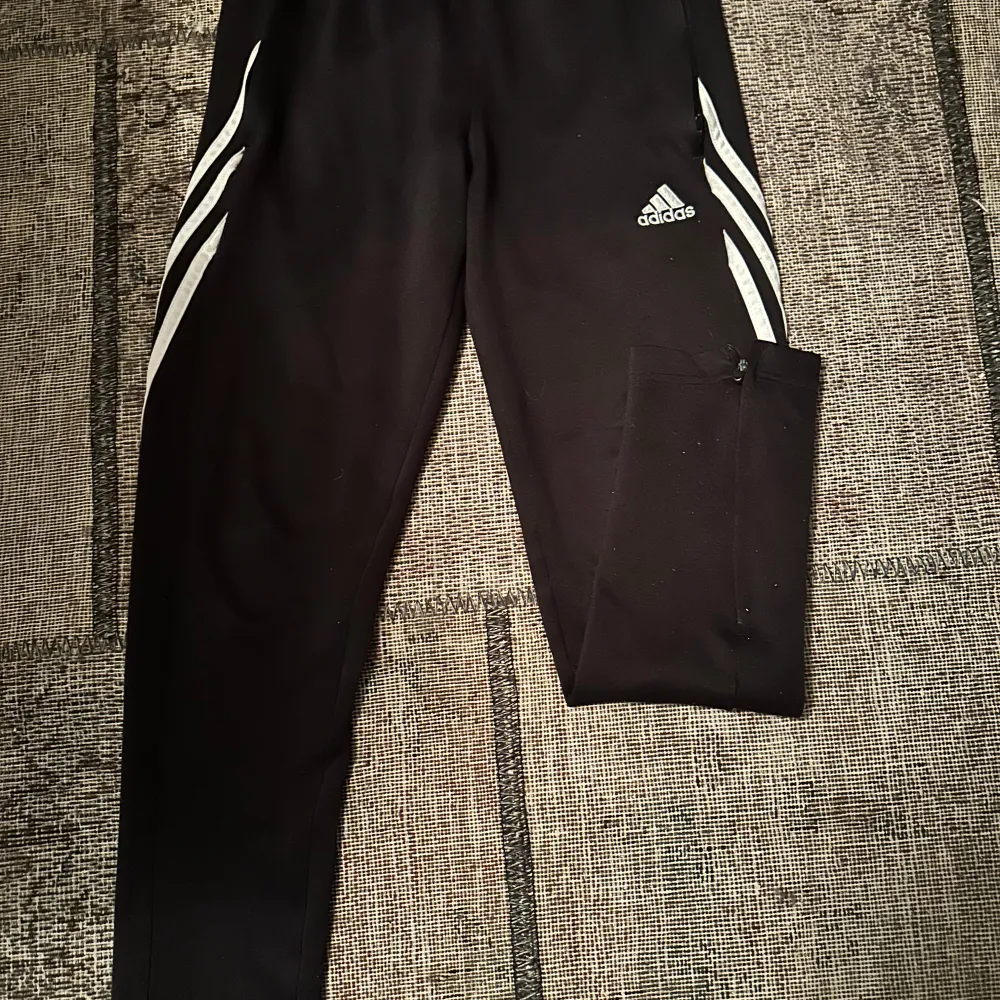 Säljer ett par svarta träningsbyxor från Adidas i storlek 152. Byxorna har de klassiska vita ränderna längs sidorna och fickor med dragkedja. Perfekta för träning eller en avslappnad dag. De är i bra skick och redo för nya äventyr!. Jeans & Byxor.