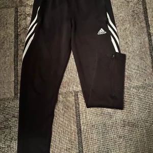 Svarta träningsbyxor från Adidas - Säljer ett par svarta träningsbyxor från Adidas i storlek 152. Byxorna har de klassiska vita ränderna längs sidorna och fickor med dragkedja. Perfekta för träning eller en avslappnad dag. De är i bra skick och redo för nya äventyr!