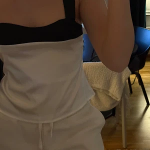 Vit och svart topp med knytband - Säljer en stilren vit topp med svarta detaljer. Toppen har breda svarta axelband och ett snyggt knytband i ryggen. Perfekt för sommardagar eller en avslappnad festkväll. Den ärmlösa designen gör den luftig och bekväm.