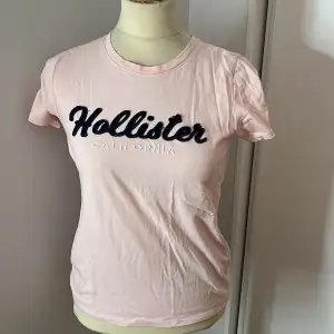 Säljer en söt rosa t-shirt från Hollister med texten 'Hollister California' i mörkblått och vitt på bröstet. T-shirten är kortärmad och i bomull, perfekt för en avslappnad stil. Passar bra till både jeans och shorts. Skicka ett meddelande om du är intresserad! 💖