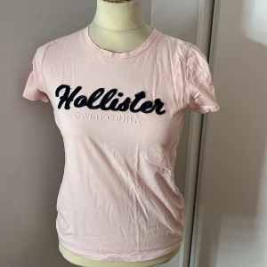 Rosa t-shirt från Hollister - Säljer en söt rosa t-shirt från Hollister med texten 'Hollister California' i mörkblått och vitt på bröstet. T-shirten är kortärmad och i bomull, perfekt för en avslappnad stil. Passar bra till både jeans och shorts. Skicka ett meddelande om du är intresserad! 💖