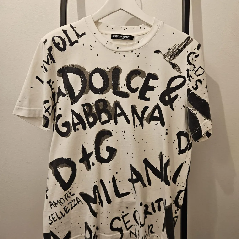 Säljer en supercool vit t-shirt från Dolce & Gabbana med svart graffiti-liknande text och mönster över hela. Den är kortärmad och perfekt för en avslappnad stil. . T-shirts.