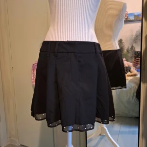 Svart kjol från Divided - Snygg svart kortkjol från Divided med knappar i framsidan och ett diskret prickigt band längst ner. Perfekt för att ge en stilren look r. Kjolen har hällor för bälte och en klassisk passform.