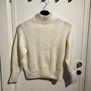 Vit stickad polotröja - Säljer en mysig vit stickad polotröja med ett klassiskt flätmönster. Tröjan har långa ärmar och ribbade muddar vid ärmslut och nederkant. Perfekt för kyliga dagar!