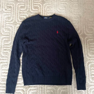 Mörkblå stickad tröja från Polo Ralph Lauren - Säljer en stilren mörkblå stickad tröja från Polo Ralph Lauren med det klassiska röda logotypbroderiet på bröstet. Tröjan har en rund hals och långa ärmar med ribbade muddar. Perfekt för en elegant och avslappnad look.