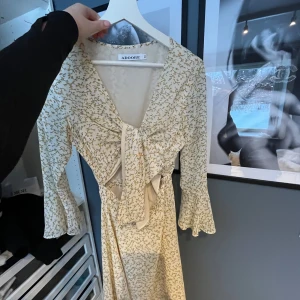 Blommig klänning från Adoore - Charmig blommig klänning från Adoore i ljus färg med små blommor. Klänningen har en omlott-design med knytning framtill och trekvartsärmar med volangdetaljer. Perfekt för en somrig look!