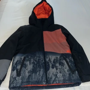 Svart/orange skidjacka från Quiksilver - Säljer en cool svart / orange skidjacka från Quiksilver med huva och dragkedja. Andvänd endast en gång och kostar 600kr i butik. Jackan har ett unikt mönster i grått och svart på nedre delen och en kontrasterande orange insida. Perfekt för kyliga dagar och skidåkning. 🎿 ⛷️☃️🧣