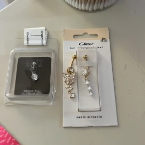 Piercingsmycken i kirurgiskt stål och 925 sterling silver - Två piercingsmycken, ett i kirurgiskt stål med cubic zirconia och ett i 925 sterling silver. Det ena smycket har en guldtonad stav med hängande stenar i olika former, inklusive en fjäril. Perfekt för att ge en glittrig touch till din stil.