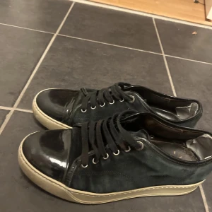 Svarta sneakers från Lanvin - Snygga svarta sneakers från Lanvin med en stilren design. Skorna har lite defekter en av de är bild 4 och sen har skosnöret på ena fått lite röd färg, vill ni ha mer information av skorna är det bara att skriva!