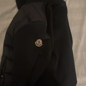 Svart jacka från Moncler - Säljer en snygg svart jacka från Moncler med huva och dragkedja. Jackan har Moncler-logga på ärmen och en cool randig detalj i rött och vitt. Perfekt för kyligare dagar. Har tyvärr inget kvitto.