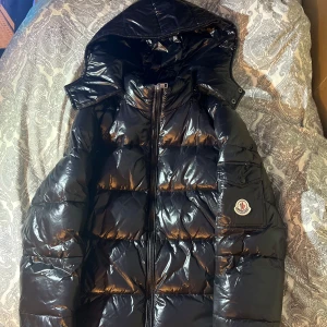 Svart pufferjacka från Moncler - Säljer en snygg svart pufferjacka från Moncler med glansig finish. Jackan har en huva och en praktisk ficka med knapp på ärmen. Perfekt för kyliga dagar med sin varma och stiliga design. Dragkedja framtill för enkel stängning.