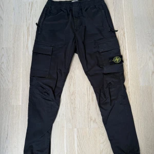 Svarta cargobyxor från Stone Island - Snygga svarta cargobyxor från Stone Island med flera praktiska fickor och elastisk midja. Byxorna har en cool patchdetalj på benet och är perfekta för en avslappnad stil.