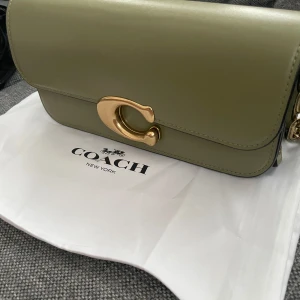 Olivgrön axelväska från COACH - Snygg olivgrön axelväska från Coach i skinn med guldfärgad kedja och spänne. Väskan har en elegant design med ett praktiskt inre fack och en justerbar rem. Perfekt för att ge din outfit en stilfull touch. Ordinarie pris 6400kr. Helt ny och oanvänd alla prislappar sitter kvar , en dustbag följer med. Skicka prisförslag .
