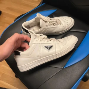 Vita sneakers från Prada - Snygga vita sneakers från Prada med klassisk design och snörning. Skorna har en diskret logga på sidan och en svart detalj vid hälen. Perfekta för en stilren look.