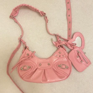 Super söt rosa axelväska med detaljer - Snygg rosa axelväska köpt i shop i USA men aldrig använd. Väskan har en justerbar axelrem och kommer med en matchande liten plånbok och hjärtformad accessoar. Hör av dig om du har några frågor :) lite starkare rosa i verkligheten 