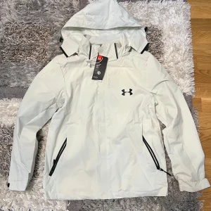 Vit vindjacka från Under Armour - Säljer en stilren vit vindjacka från Under Armour. Jackan har en justerbar huva och dragkedja framtill. Den är designad med svarta detaljer och har praktiska fickor med dragkedjor. Perfekt för blåsiga dagar.