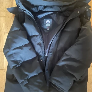 Svart dunjacka från Canada Goose - Säljer en svart dunjacka från Canada Goose, perfekt för kalla dagar. Jackan har en klassisk design med dragkedja och knappar framtill. Den är långärmad och har en hög krage för extra värme. En stilren och funktionell jacka för vintern.