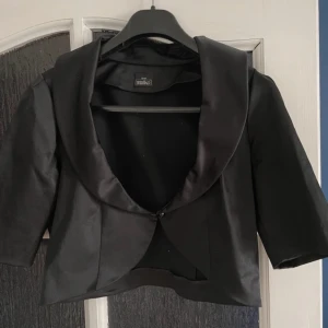 Jättefin bolero - Elegant svart kavaj från Salko med trekvartsärm och en knapp framtill. Den har en glansig finish som ger en lyxig känsla. Perfekt för att lyfta din outfit till nästa nivå.