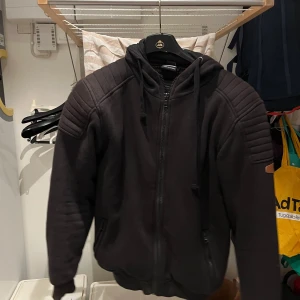 Svart jacka med huva - Säljer en svart jacka med huva och axel och arm skydd. Jackan har quiltade detaljer på axlarna och dragkedja framtill. Perfekt för att åka moped eller mc 