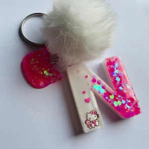 Resin Nyckelring med bokstaven N och Hello Kitty - Söt nyckelring med bokstaven N i rosa glitter och en vit fluffig boll. Dekorerad med en liten Hello Kitty-figur och rosa hjärtan. Perfekt accessoar för att ge en personlig touch till dina nycklar eller väska.