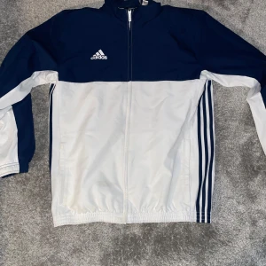 Blå och vit träningsjacka från Adidas - Säljer en snygg blå och vit träningsjacka från Adidas. Jackan har dragkedja framtill och de klassiska tre ränderna längs sidorna. Perfekt för sportiga aktiviteter eller en avslappnad stil. Adidas-loggan är broderad på bröstet.