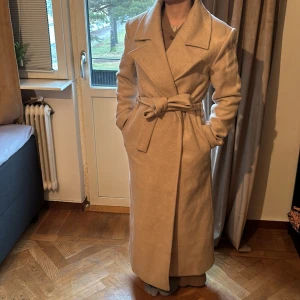 Beige kappa från NA-KD - Säljer en elegant beige kappa från NA-KD. Den har en klassisk design med knytbälte i midjan och långa ärmar. Perfekt för kyligare dagar med sin långa längd och stilrena look. För lång på mig som är 1.58 lång🖤