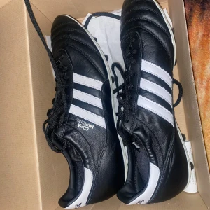 Adidas Copa Mundial fotbollsskor - Säljer ett par klassiska Adidas Copa Mundial fotbollsskor i svart och vitt. Skorna har snörning och är tillverkade i skinn med dobbar för optimalt grepp på planen. Perfekta för fotbollsentusiaster som vill ha både stil och funktion.