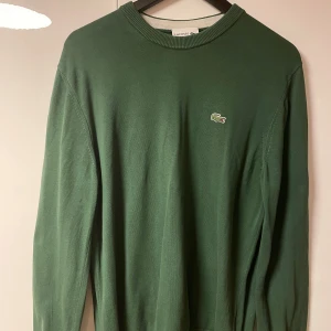Grön tröja från Lacoste - Säljer en stilren grön tröja från Lacoste med det klassiska krokodilemblem på bröstet. Tröjan har en normal passform och är långärmad, perfekt för en avslappnad look. 🐊 Tröjan har en liten vit linje vid ärmen, vilket verkar vara en del av designen. Den är varsamt använd och ser ut som ny. Hör av dig vid frågor!