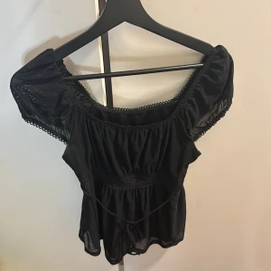 Svart offshoulder blus - Snygg svart offshoulder blus med korta ärmar och spetsdetaljer. Blusen har en luftig passform och knytband i midjan för en feminin look. Perfekt för en stilren outfit. Den är från SHEIN