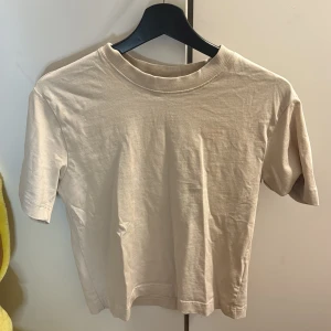 Beige t-shirt från Gina Tricot - Säljer en stilren beige t-shirt från Gina Tricot. Den har en klassisk rund hals och korta ärmar, perfekt för en enkel och avslappnad look. Gjord i mjukt material för extra komfort. Behöver bara strykas lite , sen blir den perfekt!