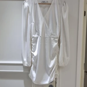 Vit klänning  NA-KD - Säljer en elegant vit klänning från NA-KD med långa ärmar och v-ringning. Klänningen har en stilren design med draperade detaljer på sidorna som ger en snygg passform. Perfekt för en stilfull look.