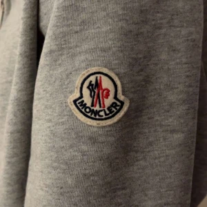 Grå huvtröja från Moncler - Säljer en stilren grå huvtröja från Moncler med deras ikoniska logga på ärmen. Tröjan har en huva med ränder i rött, vitt och blått. Perfekt för en casual look.