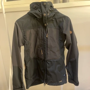 Svart jacka från Fjällräven - Säljer en svart jacka från Fjällräven i storlek XS. Jackan har en huva och dragkedja framtill med knappar. Perfekt för utomhusaktiviteter med sin funktionella design och slitstarka material. Älskar den och så man är så rörlig i den men säljer då ja växt ur den :( 