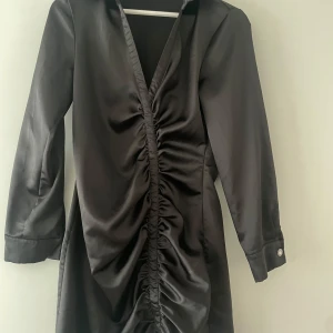 Svart omlottklänning med knappar - Svart omlottklänning i satin med långa ärmar och v-ringad design. 