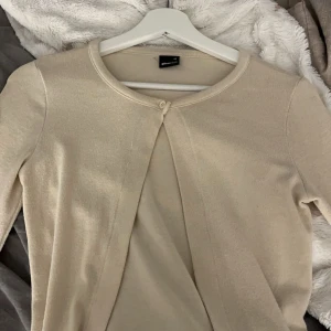 Beige kofta från Gina Tricot - Säljer en stilren beige kofta från Gina Tricot i storlek XS. Koftan har en enkel knapp framtill och är perfekt för lager-på-lager-stil. Den är långärmad och gjord i ett mjukt material som känns skönt mot huden.