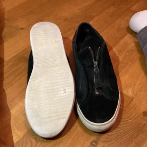 Arigato - Snygga svarta sneakers med dragkedja framtill. Skorna har en vit sula och är tillverkade i mocka och skinn. Perfekta för en stilren look. De är slitna men jag har tvättat väldigt bra och skick 6/10. Meddela gärna. Ett tips lägg i ett till skoinlägg om de är lite stora.Det är trasiga i sömmarna men och tråden har gått upp lite som på bilden. Väldigt stora för sin storlek 
