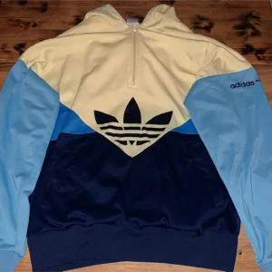 Flerfärgad retrohoodie från Adidas - Säljer en cool flerfärgad hoodie från Adidas med dragkedja och stor logga på i sammet på bröstet. Tröjan har en kombination av ljusgul, ljusblå och mörkblå färger. Perfekt för en avslappnad stil.