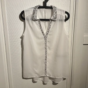 Vit ärmlös blus med pärldetaljer - Snygg vit ärmlös blus från vero moda med pärldetaljer längs kragen och knäppningen. Perfekt för en stilren look med en touch av glamour. Blusen har en lätt och luftig design.